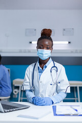 Hospital medical specialist sitting at office table while wearing virus protection facemask and surgical gloves. Clinic healthcare expert sitting at office desk while wearing facemask