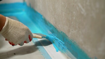 A worker is applying waterproofing paint to the floor in the bathroom. The master with a brush...