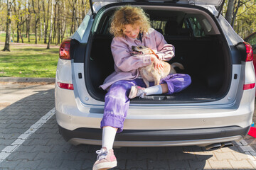 Curly-haired beautfiul attractive girl in her 20s hugging her small chunky dog in a car trunk on a parking lot, sitting with her legs crossed. High quality photo - Powered by Adobe