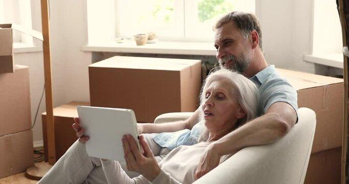 Happy older wife relax on comfy couch at moving day lean back on beloved aged husband hold touchpad discuss future living room interior with spouse. Mature couple realty buyers use tablet at new home