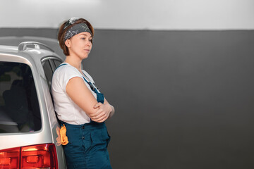 Caucasian young woman in a blue coveralls leaning at the car. Copy space. Concept of work in an auto repair shop