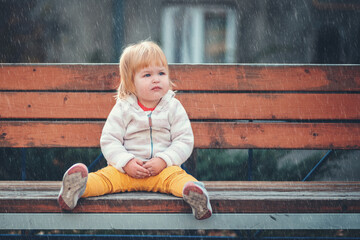 Childrens shelter. A sad little girl is sitting on a bench on the playground alone. Rainy weather. The concept of lost childrens