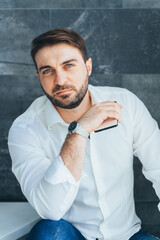 Vertical image of handsome unshaven businessman, sitting indoors in loft. Holding smartphone in hands. Look camera