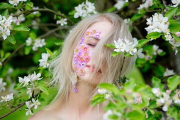 Fototapeta premium Beautiful woman with flowers petals on face around blooming apple tree