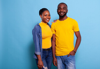 Fototapeta premium Smiling couple standing in embrace over blue background. Man and woman in love expressing love and affection with hug in studio. Lovers feeling happy and peaceful, looking at camera.