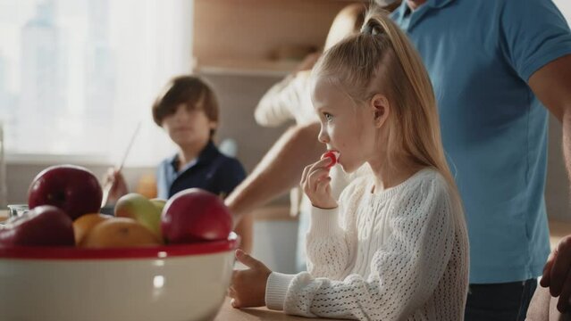 Close-up shooting of nice little Caucasian girl with ponytail holding tomato in hand and eating vegetable. Father, mother and brother on background. Healthy food. Eco trend. Kitchen, home, apartment
