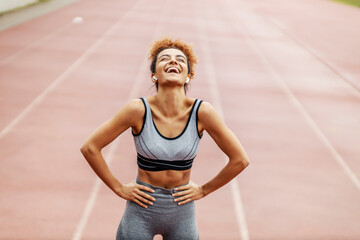 A happy runner stands in the stadium with her hands on hips and laughing. She is so happy she...