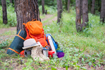 Hiking equipment in a pine forest. Backpack, thermos, sleeping bag, compass, hat and shoes