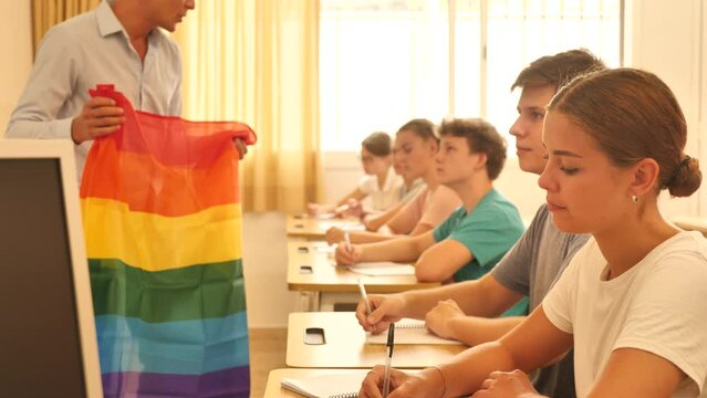 Focused teenage schoolgirl sitting at lesson with classmates, listening young teacher talking about LGBT community and showing rainbow flag. High quality 4k footage