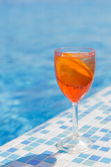 A transparent glass with an orange citrus cocktail stands on the edge of the swimming pool