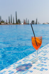 Orange cocktail with a straw on the background of the pool with clear blue water, in the background of the park