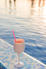 Orange cocktail in a glass with a straw on the background of blue water in the pool with a reflection of the sunset