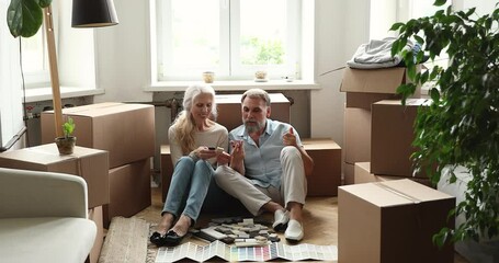 Inspired older adult husband wife engaged in apartment renovation sit at living room full of packed staff choosing decor. Senior couple plan home redecoration select tile using samples paper catalogue