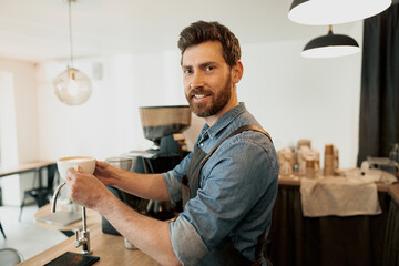 Handsome barista holding cup of coffee and look at the camera standing behind a counter - obrazy, fototapety, plakaty