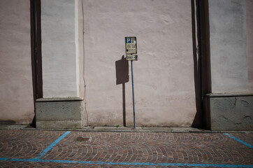Old sign of paid parking by time and pointer to parking ticket in Italy. Parking space with blue markings on paved street and pointer to parking machine against pink wall in old town in Borgo Taro