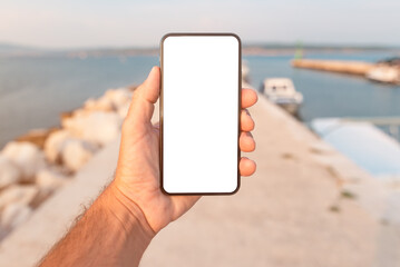 Man standing by the sea and holding mobile smart phone with blank mockup screen, closeup of hand