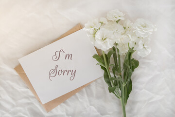 a white small bouquet on a white sheet, a craft envelope and a card with the inscription I am sorry