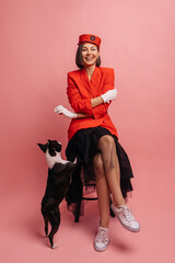 Stylish young caucasian woman playing with little bulldog on pink background. Laughing brunette wears red jacket and black skirt. True friendship, wonderful happy moments