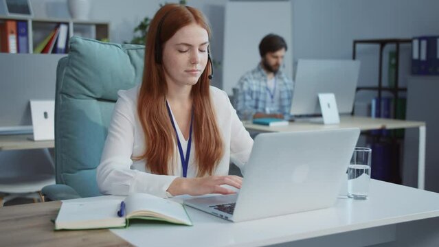 Footage of young beautiful redhead woman with long hair in formal clothes sitting at silver laptop, talking to client. Female call operator using computer and headset for consulting clients online.