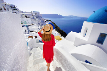 Beautiful girl in red dress and hat comes down the stairs exploring Oia village with spectacular view of the Caldera, Santorini, Greece - obrazy, fototapety, plakaty
