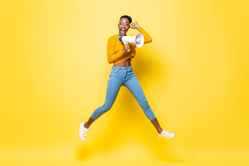 Fototapeta na wymiar Portrait of cheerful young African American woman holding megaphone jumping in isolated yellow background