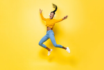 Full body portrait of happy African American woman listening to music and jumping with hands raising in yellow isolated studio color background