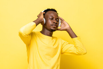 Fototapeta na wymiar Young African man listening to music with eyes closed while holding headphones against yellow studio background