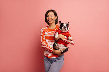 European young woman tenderly holds french bulldog in hoodie on pink background. Brunette girl...