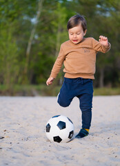 Kid Kicks Football Ball. Funny Little Boy Playing with Black and White Soccer Ball in Park on Sand