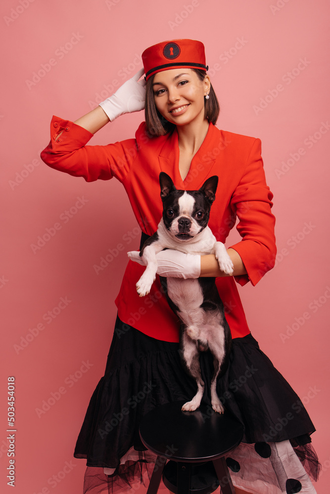 Wall mural Beautiful young caucasian woman holding purebred dog, feeling happy on pink background. Brunette girl in red suit looks at camera. Animal owner concept - Wall murals