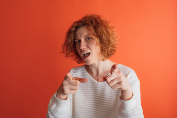 Portrait of red-haired woman pointing with fingers at camera isolated over red studio background