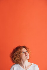 Dreaming. Portrait of emotive red-haired woman with curly hair making funny thoughtful expression isolated over red studio background.