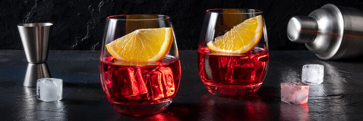 Campari or negroni cocktails on a black slate background with fresh oranges, a jigger, and a...