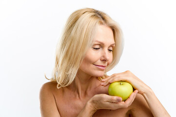 Calm senior woman holding fresh green apple middle her hands and smiling. Elderly model with naked shoulders posing and looking at the fruit isolated on white. Anti age treatment