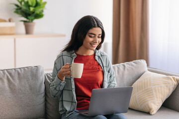 Happy Arab businesswoman having online business meeting on laptop, drinking coffee at home office