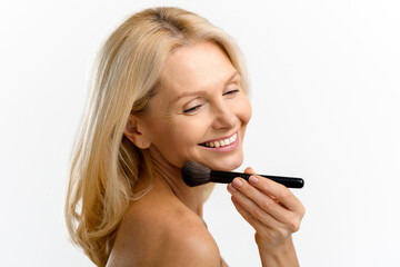 Blonde 40s 50s middle aged woman with holding make up brush on shining face with perfect skin, looking away, applying powder or some cosmetic product. Advertising concept