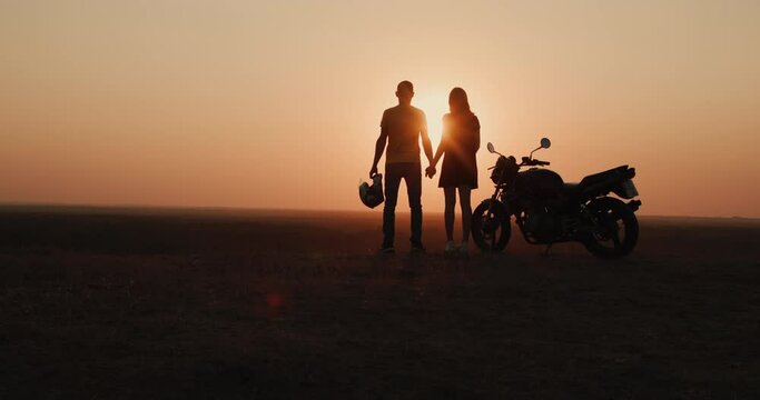 Silhouettes of a young couple at sunset, standing at the motorcycle looking at the setting sun