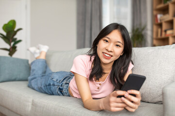 Portrait of positive asian lady using smartphone to work or study, lying on sofa at home and smiling at camera