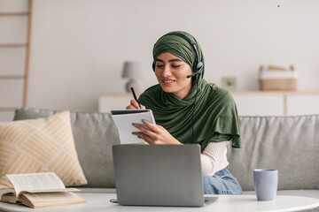 Cheerful Arab woman in hijab using laptop, wearing headphones, writing down information during...