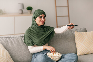 Happy young Arab woman with remote control sitting on couch, watching TV and eating popcorn at home