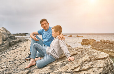 Happy family, father and son bonding, sitting on stone by the sea looking at view enjoying summer vacation. Togetherness Friendly concept