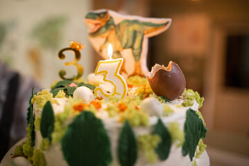 Cake with a picture of a dinosaur egg and a tyrannosaurus rex in the background. candle number 3