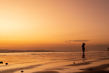 Fototapeta premium Woman enjoying life on the beach at sunset. Silhouette of young woman standing feeling free and watching golden hour beautiful colors