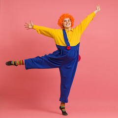 a cheerful clown in a wig and a yellow-blue suit jumps on one leg on a colored background