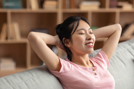 Rest And Relax In Your Own Home. Happy Asian Lady With Closed Eyes Threw Hands Behind Head Sitting On Sofa