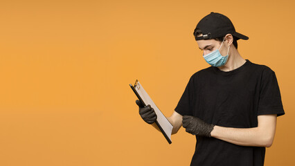 delivery man in a black t-shirt, mask and gloves looks at documents on a clipboard, yellow background with copy space