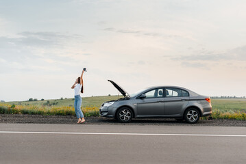Fototapeta na wymiar A young girl stands near a broken car in the middle of the highway during sunset and tries to call for help on the phone and start the car. Waiting for help. Car service. Car breakdown on the road.