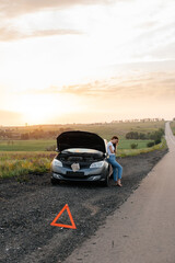 Obraz na płótnie Canvas A frustrated young girl stands near a broken-down car in the middle of the highway during sunset. Breakdown and repair of the car. Waiting for help. Car service. Car breakdown on road.