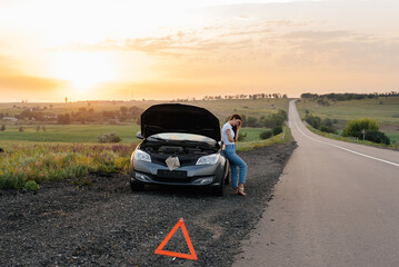 A frustrated young girl stands near a broken-down car in the middle of the highway during sunset. Breakdown and repair of the car. Waiting for help. Car service. Car breakdown on road.