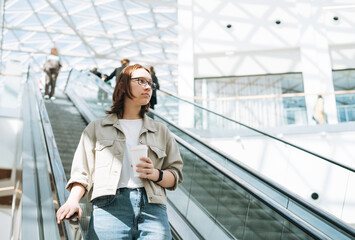 Obraz premium Young brunette teenager girl student in glasses on escalator in shopping mall public place